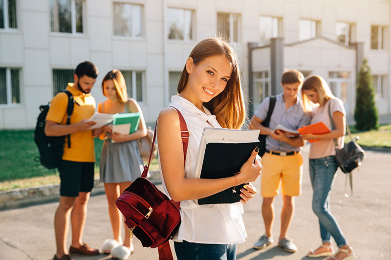 Handsome young girl with red velvet backpack holding books and smiling while standing against university with her friends in the background
